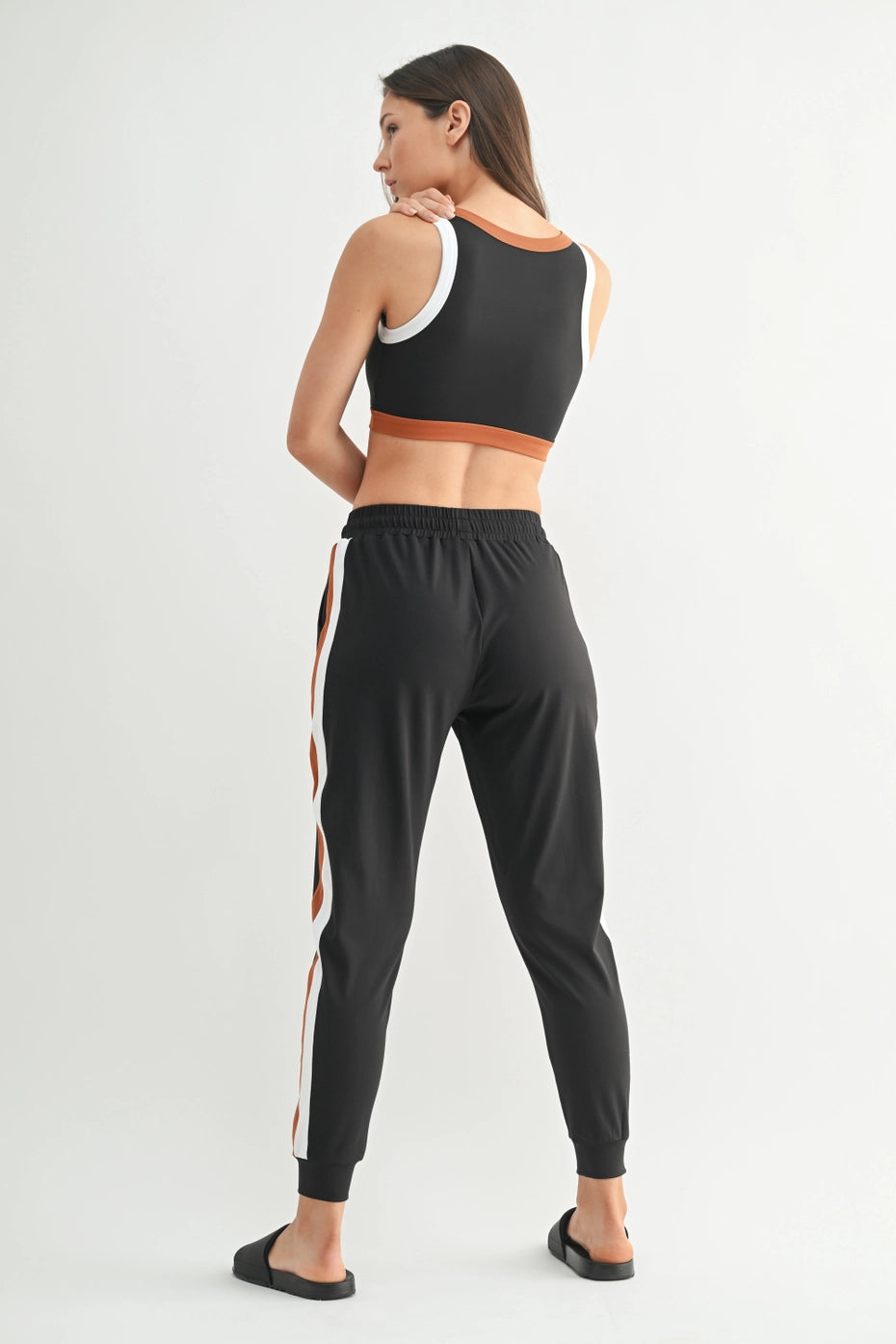 Backside profile Woman wearing a black sports bra and black jogger pants with white and  orange stripes on a white background